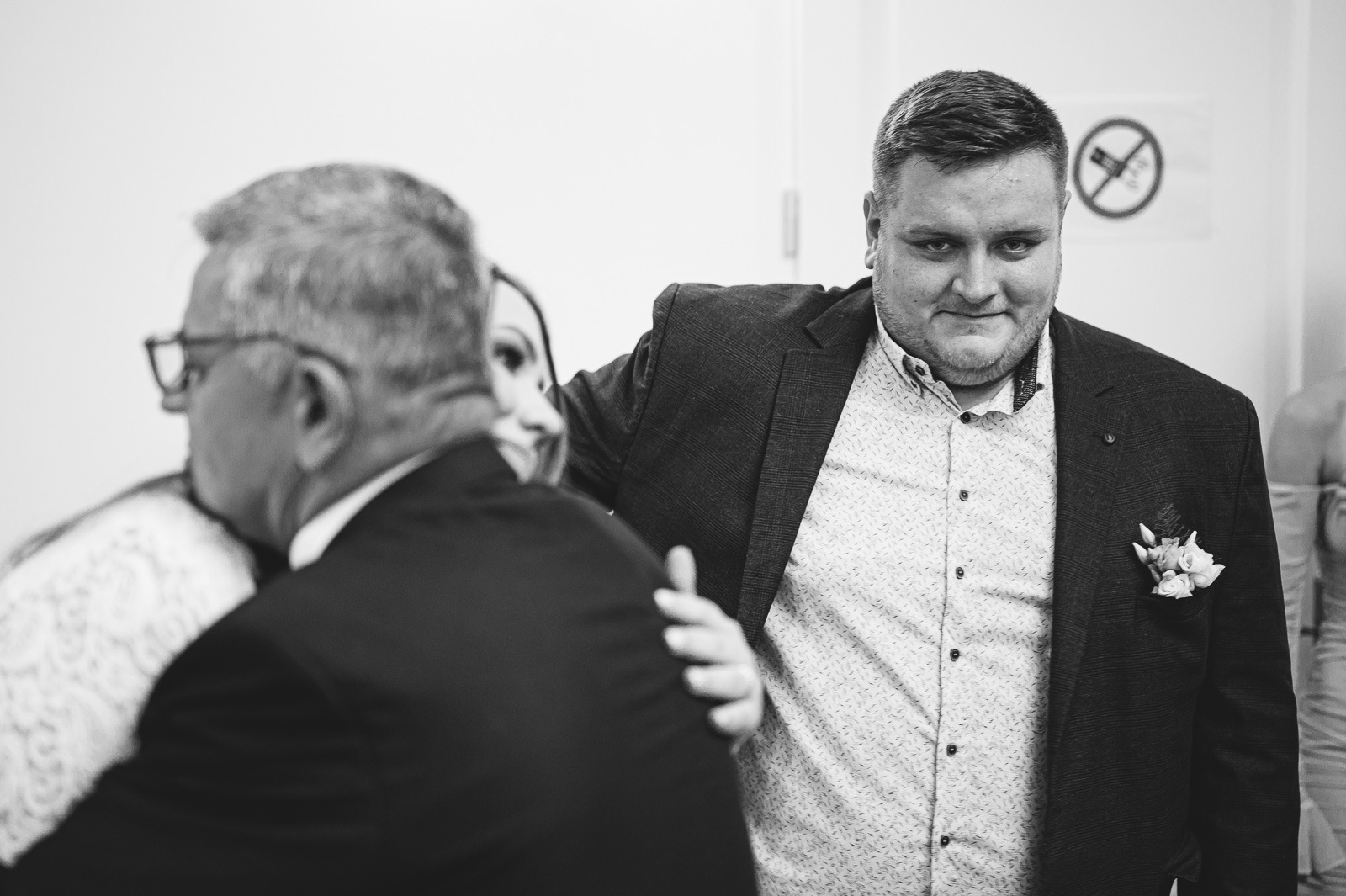 Czarno-białe zdjęcie z reportażu ślubnego: mężczyzna w garniturze z butonierką obejmuje parę młodą, na ścianie znak zakazu palenia.