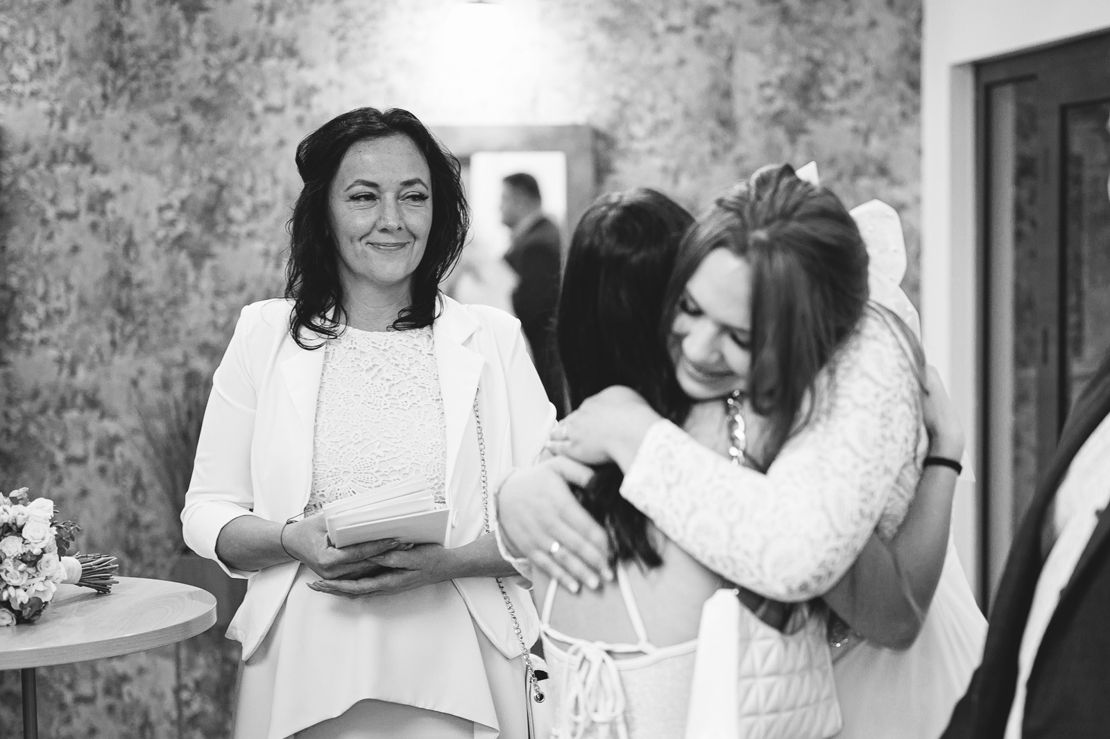 Czarno-białe zdjęcie z reportażu ślubnego: uśmiechnięta kobieta w białym żakiecie trzymająca zaproszenia, obok panna młoda obejmująca gościa, w tle lustro z odbiciem mężczyzny.