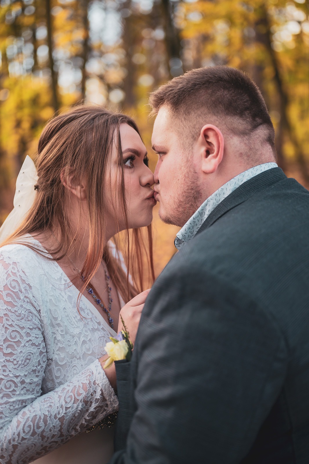Pocałunek pary młodej w jesiennej scenerii, panna młoda w koronkowej sukni z bukiecikiem, pan młody w ciemnym garniturze.