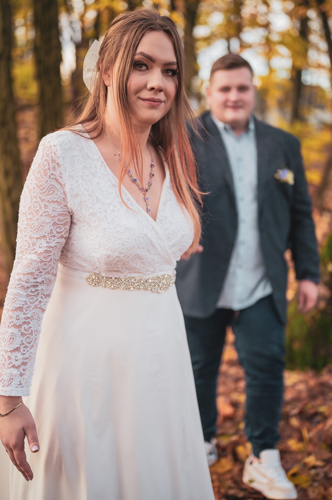 Uśmiechnięta panna młoda w koronkowej sukni z ozdobnym pasem, w tle zamazany pan młody w garniturze, jesienna sceneria.