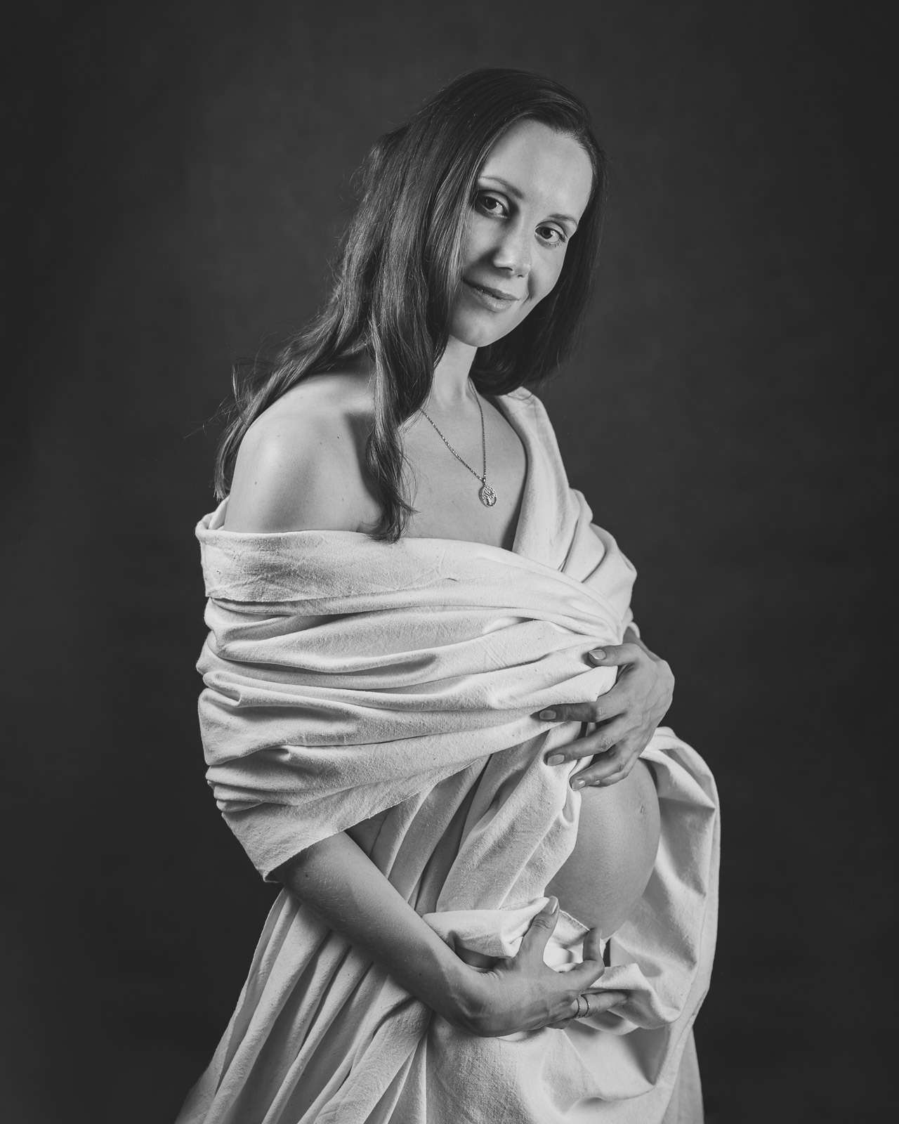 Czarno-białe portretowe ujęcie kobiety w ciąży, owiniętej w białą tkaninę, z delikatnym uśmiechem i naszyjnikiem z symbolem drzewa życia na szyi, na ciemnym tle.