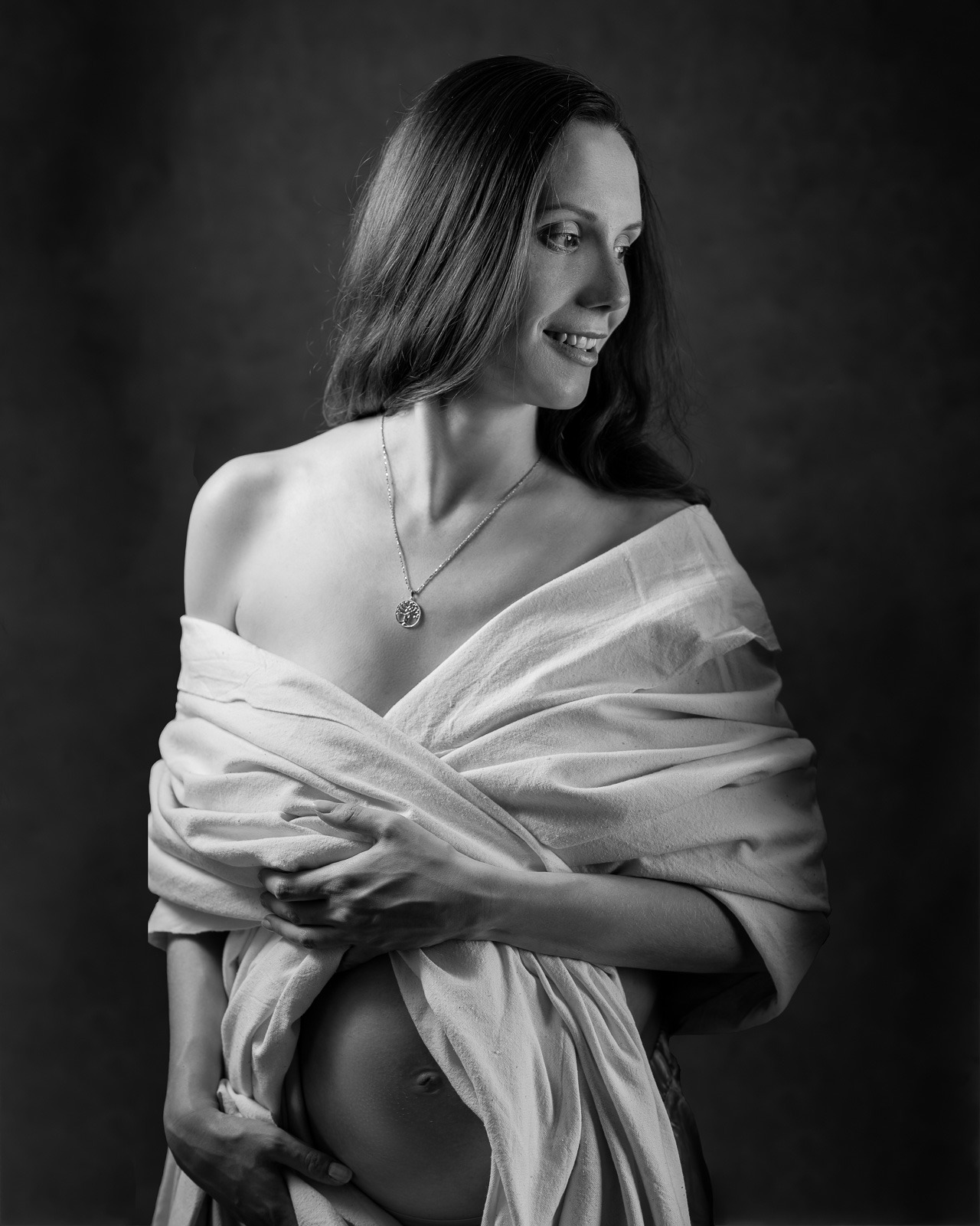 Czarno-biała fotografia portretowa uśmiechniętej, szczupłej kobiety w ciąży, owiniętej w białą tkaninę, z delikatnym naszyjnikiem z motywem drzewa życia, na ciemnym tle.