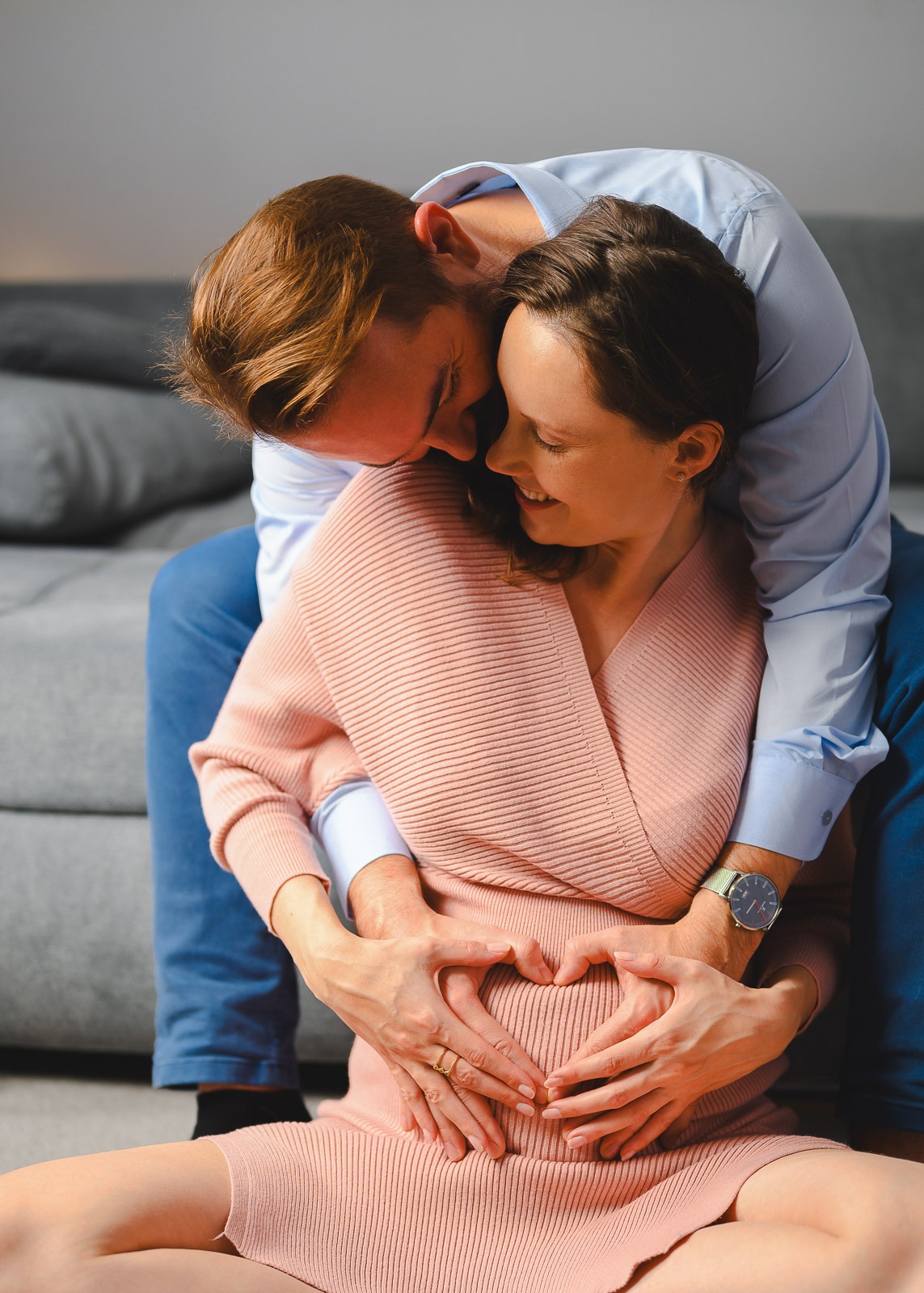 Przyszły tata obejmuje uśmiechniętą kobietę w różowej sukience, dłonie na brzuchu ułożone w kształt serca, widoczny zegarek na ręce mężczyzny i obrączka na palcu kobiety.