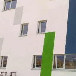 Fasada budynku z oknami, pomalowana w geometryczne wzory w kolorach: biały, szary, zielony, niebieski i brązowy. Widoczne rusztowania przed drzwiami.