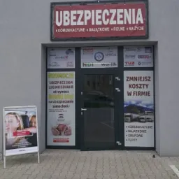 Fasada budynku z szyldem 'Ubezpieczenia' i naklejkami reklamowymi różnych towarzystw ubezpieczeniowych na drzwiach wejściowych. Na chodniku stoi tablica reklamowa z ofertą ubezpieczeń komunikacyjnych.