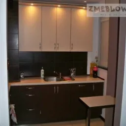 Niewielka kuchnia z brązowymi i beżowymi szafkami, czarnym backsplashiem i blatem w kolorze kremowym, oświetlona halogenami, z małym stolikiem z boku.