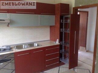 Częściowy widok kuchni z zabudową w odcieniach brązu, szafki górne z matowym szkłem, wysuwana szafka cargo, jasny blat i zlewozmywak.