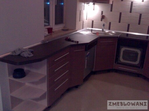 Kąt ujęcia na nową, brązową zabudowę kuchenną z ciemnym blatem, widoczne płytki na ścianie i okno w tle.