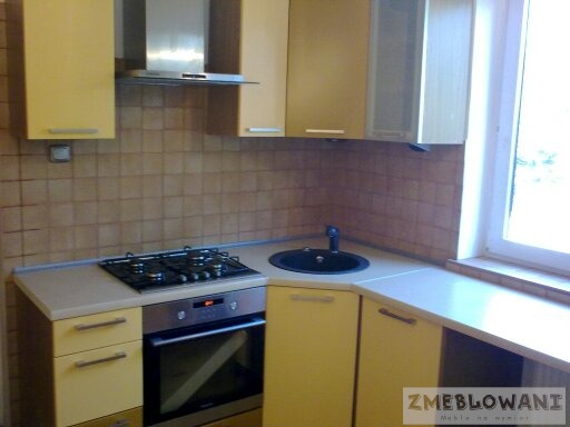 Żółta, narożna zabudowa kuchenna z piekarnikiem, płytą gazową i czarnym zlewozmywakiem, widok na fragment ściany z brązowymi płytkami.