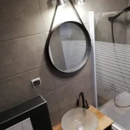 Nowoczesna łazienka z umywalką ze szkła na drewnianej szafce, okrągłym lustrem z oświetleniem punktowym i kabiną prysznicową z poziomymi paskami na szkle.