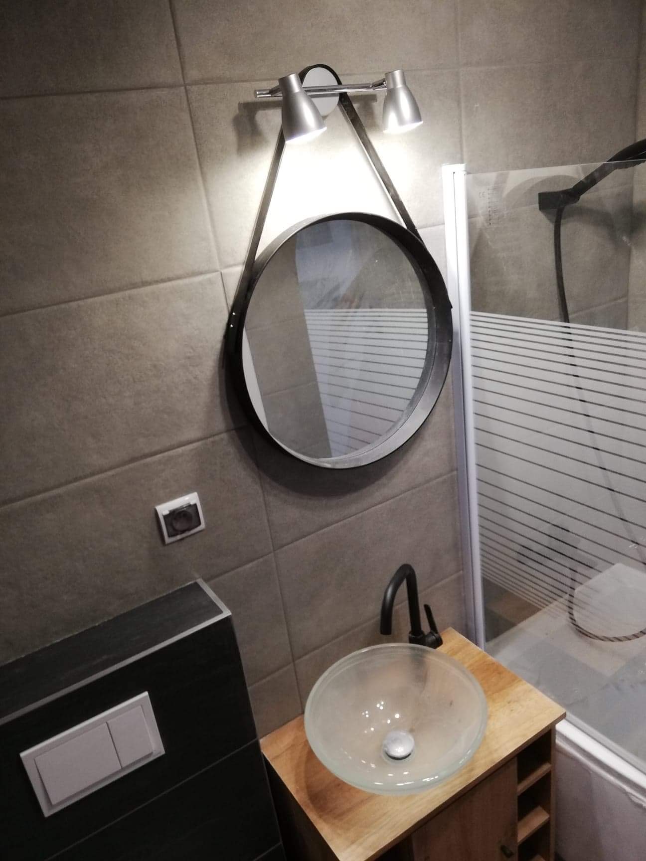 Nowoczesna łazienka z umywalką ze szkła na drewnianej szafce, okrągłym lustrem z oświetleniem punktowym i kabiną prysznicową z poziomymi paskami na szkle.