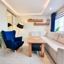 Jasne wnętrze salonu z narożnikiem, drewnianym stołem, niebieskim fotelem typu uszak i oświetleniem LED na suficie.