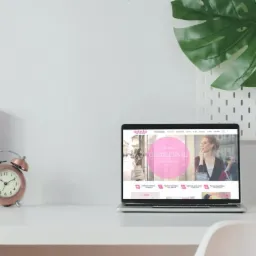 Laptop wyświetla stronę internetową 'Natasha' na minimalistycznym biurku z zegarem, rośliną i akcesoriami.