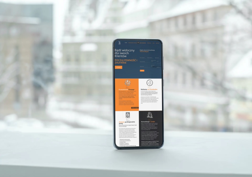 Smartfon wyświetlający stronę internetową z ofertą usług marketingowych, takich jak prowadzenie fanpage'a i reklama na Facebooku, na rozmytym tle okna i budynku.