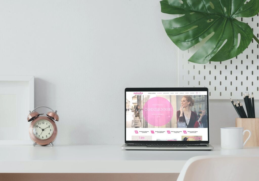Laptop wyświetla stronę internetową 'Natasha' na minimalistycznym biurku z zegarem, rośliną i akcesoriami.