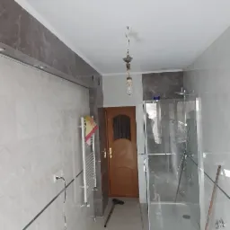 Wnętrze łazienki w trakcie remontu, widoczne płytki ścienne z geometrycznym wzorem, szklana kabina prysznicowa, grzejnik drabinkowy z powieszonym ręcznikiem, niedokończone oświetlenie sufitowe...