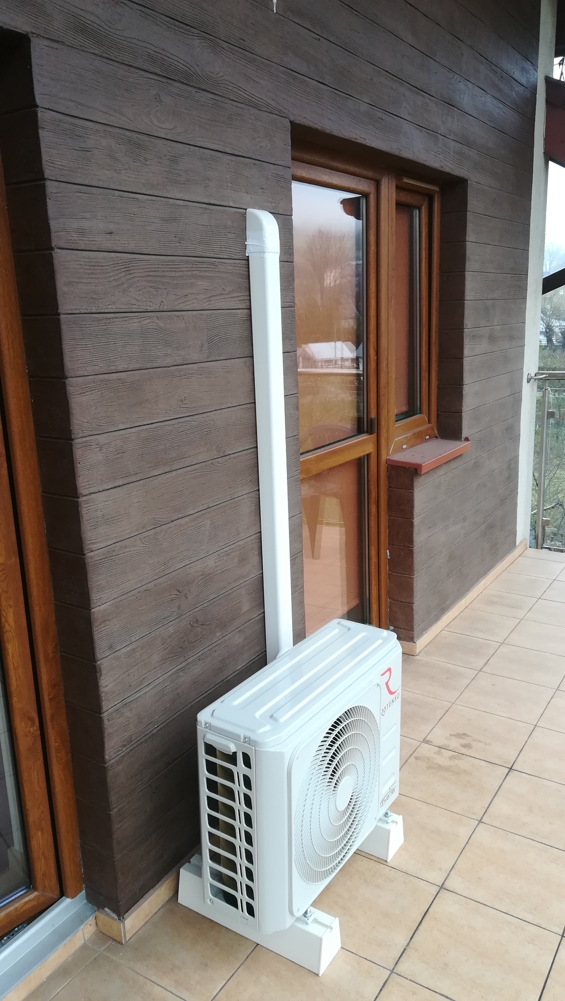 Zewnętrzna jednostka klimatyzacyjna marki Rotenso zamontowana na balkonie z brązową elewacją imitującą drewno, widoczna rura odprowadzająca skropliny.
