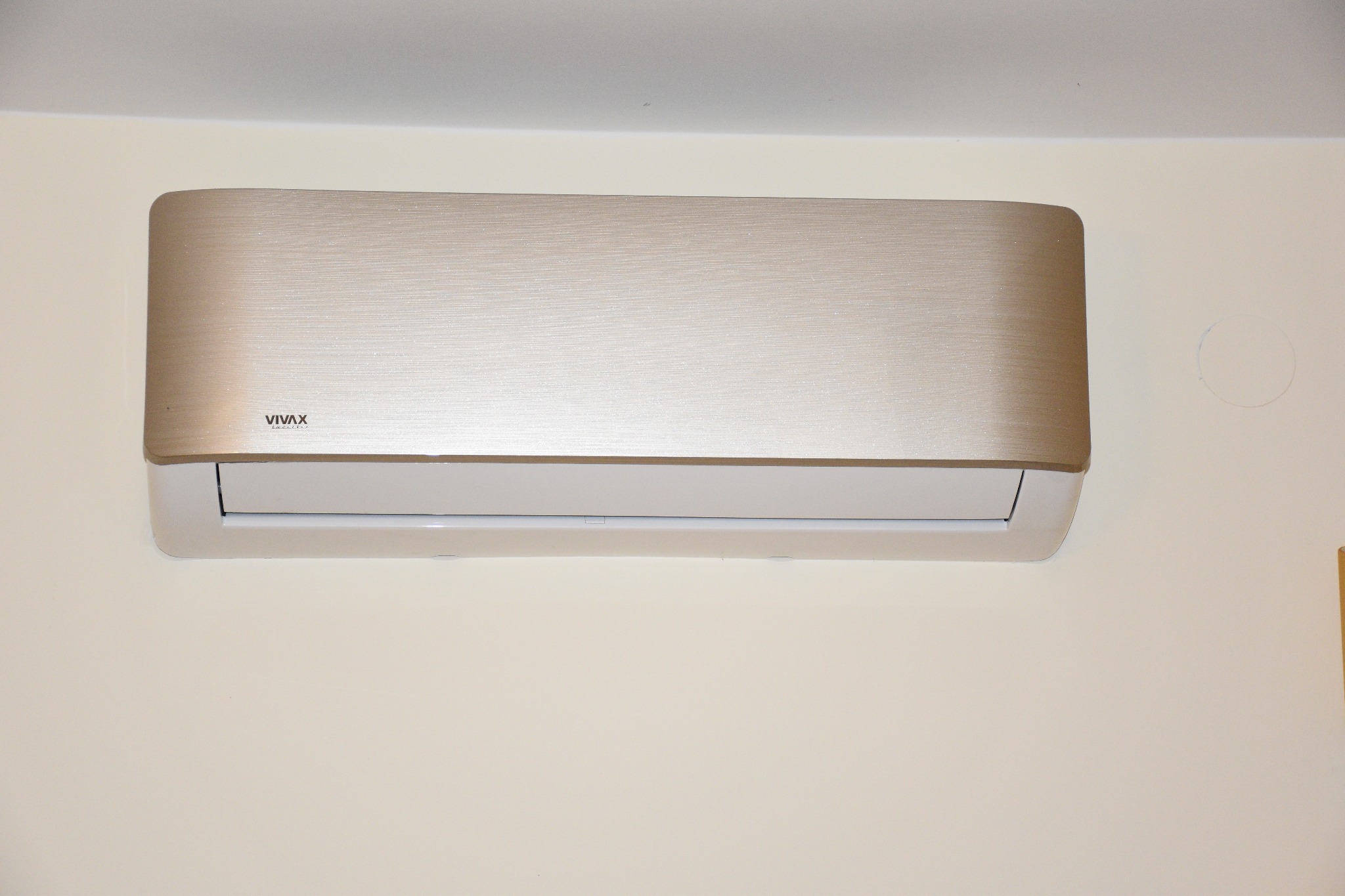 Nowoczesny, złoty klimatyzator marki Vivax zamontowany na kremowej ścianie w pomieszczeniu.