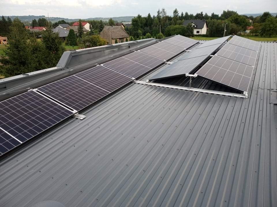 Panele słoneczne zamontowane na metalowym dachu z widokiem na wiejski krajobraz, lekko pochmurne niebo.