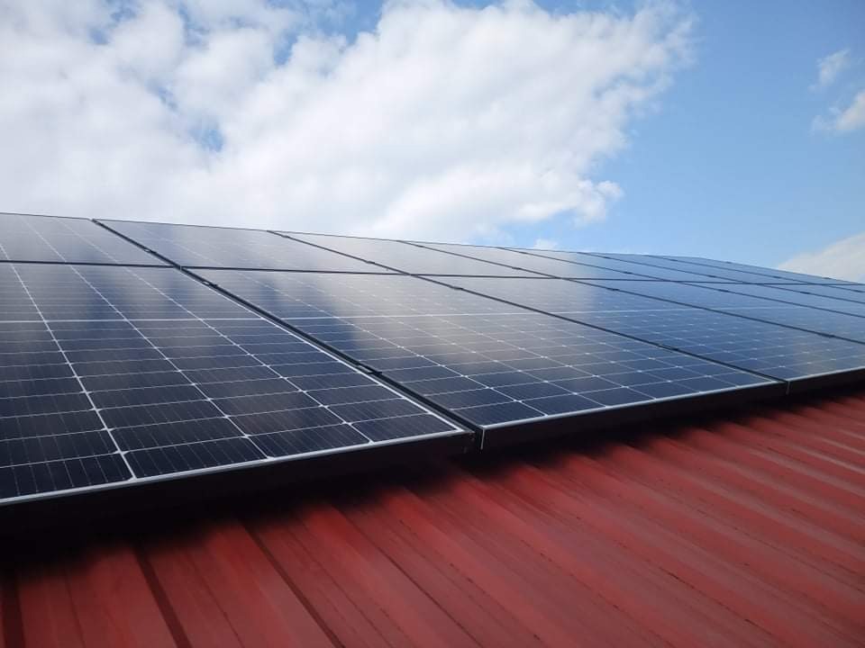 Panele fotowoltaiczne zamontowane na czerwonym, metalowym dachu, z odbiciem błękitnego nieba i chmur na powierzchni paneli.