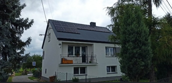 Dwupiętrowy dom z ciemnym dachem i panelami słonecznymi w Kielcach, widoczny zza drzew.