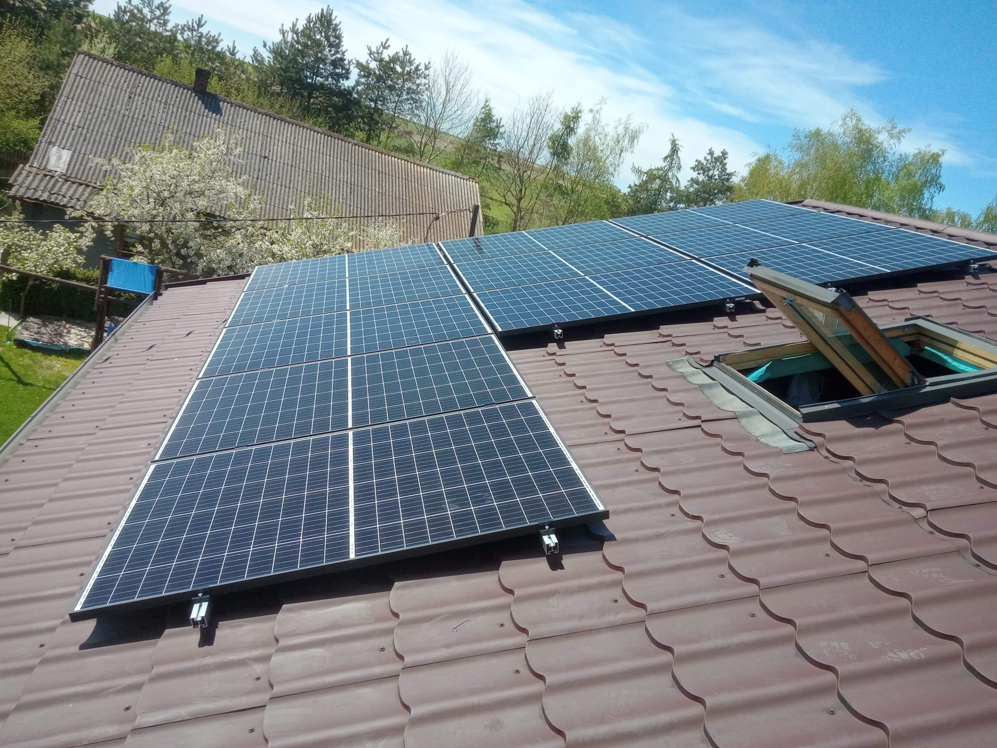 Panele słoneczne na brązowym dachu z blachodachówki, obok otwarte okno dachowe, w tle drzewa i budynki.