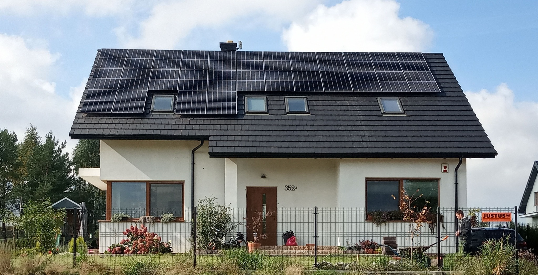 Dom jednorodzinny z panelami fotowoltaicznymi na dachu, widoczne okna dachowe, zadbany ogród z kwiatami i ogrodzeniem.