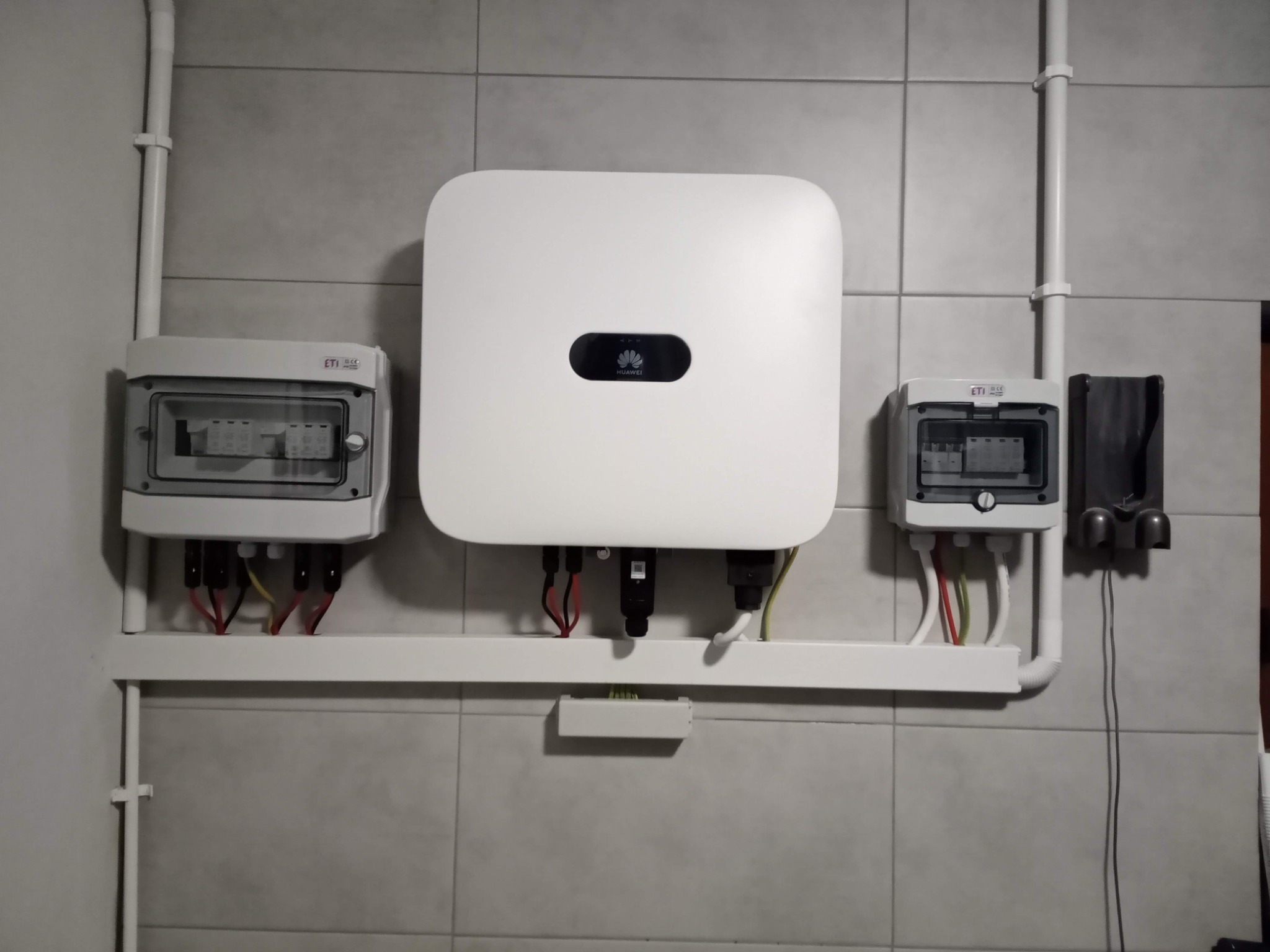 Biały inwerter Huawei zamontowany na szarej, kafelkowanej ścianie, z widocznymi przewodami i dwiema skrzynkami rozdzielczymi po bokach, całość przymocowana do ściany za pomocą białej listwy.