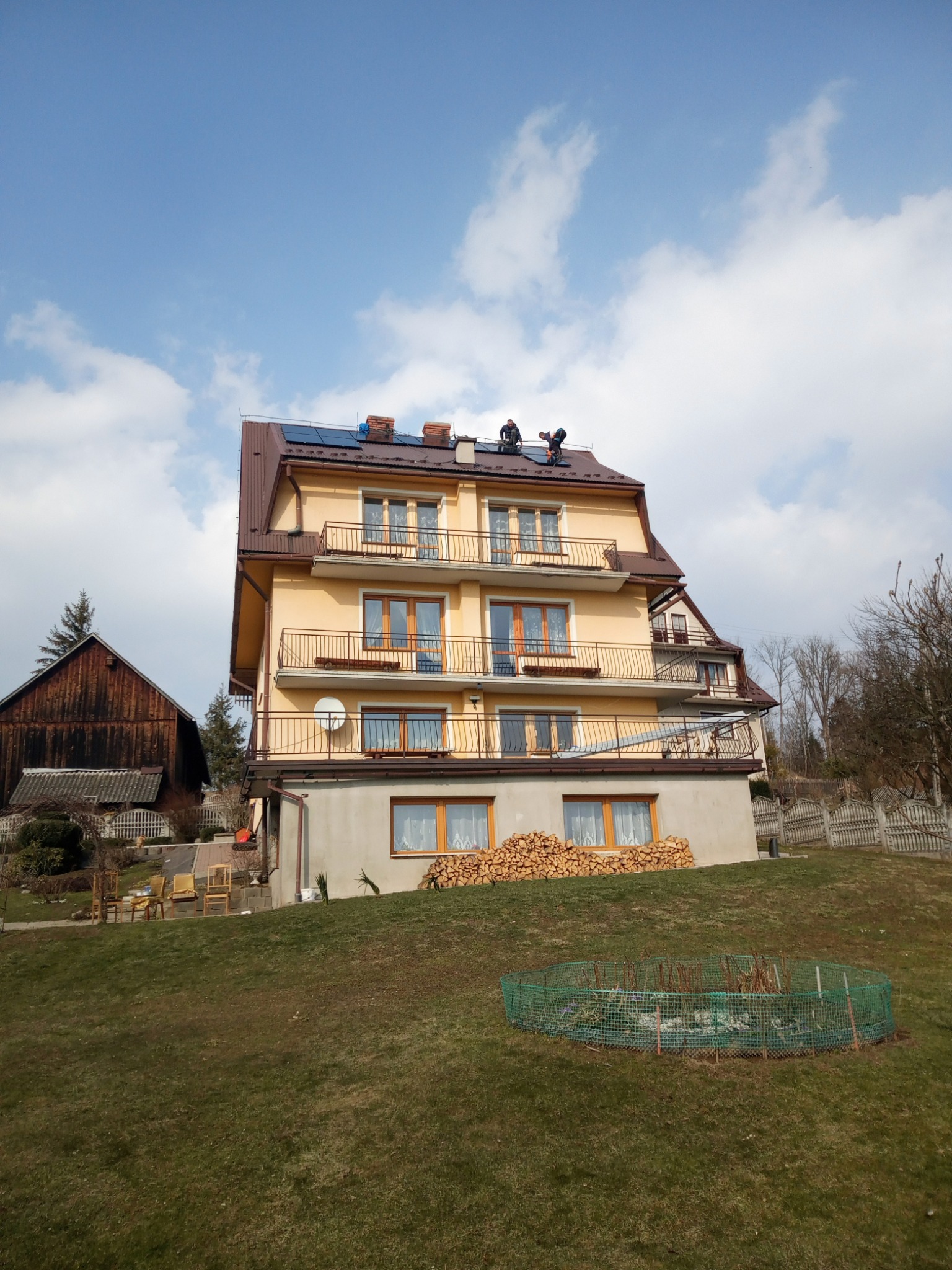 Dwóch monterów instaluje panele słoneczne na dachu dwupiętrowego domu z balkonami i stertą drewna obok, widok z dołu.