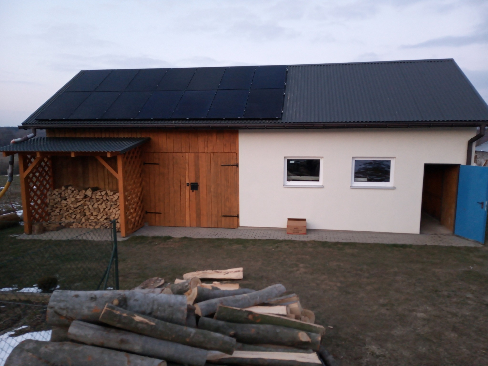 Instalacja paneli słonecznych na dachu budynku gospodarczego w Kielcach, z widocznym składem drewna opałowego i stertą pociętych pni na pierwszym planie.