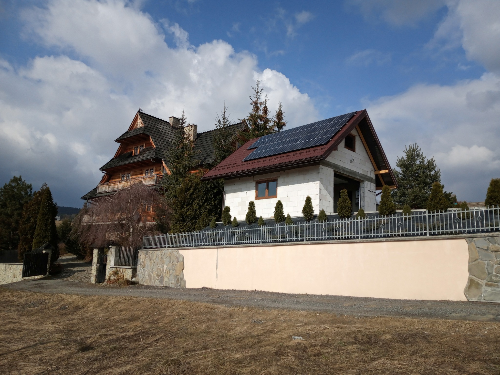 Nowoczesny dom z panelami słonecznymi na dachu, widoczny z perspektywy ulicy, obok tradycyjny góralski dom z ciemnego drewna i kamienny mur.