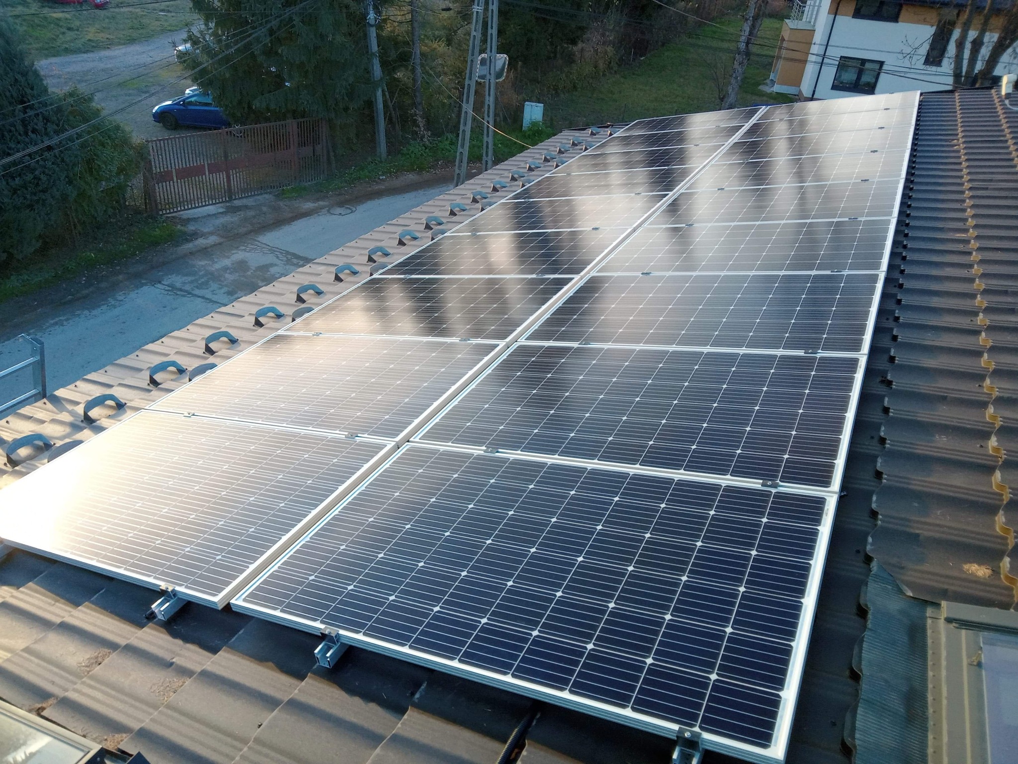 Panele słoneczne zamontowane na ciemnym dachu z dachówki ceramicznej, widok z góry na instalację z widocznymi elementami mocującymi i otoczeniem budynku.