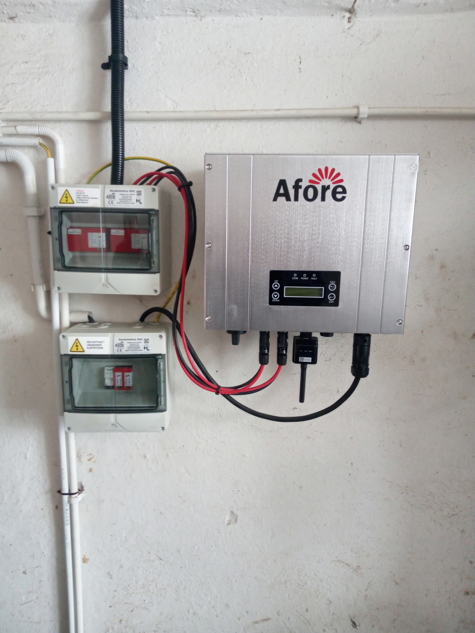 Srebrny inwerter Afore zamontowany na pomalowanej, nierównej ścianie, podłączony do dwóch rozdzielnic z bezpiecznikami w przezroczystych obudowach. Widoczne przewody elektryczne w kolorach czarnym...