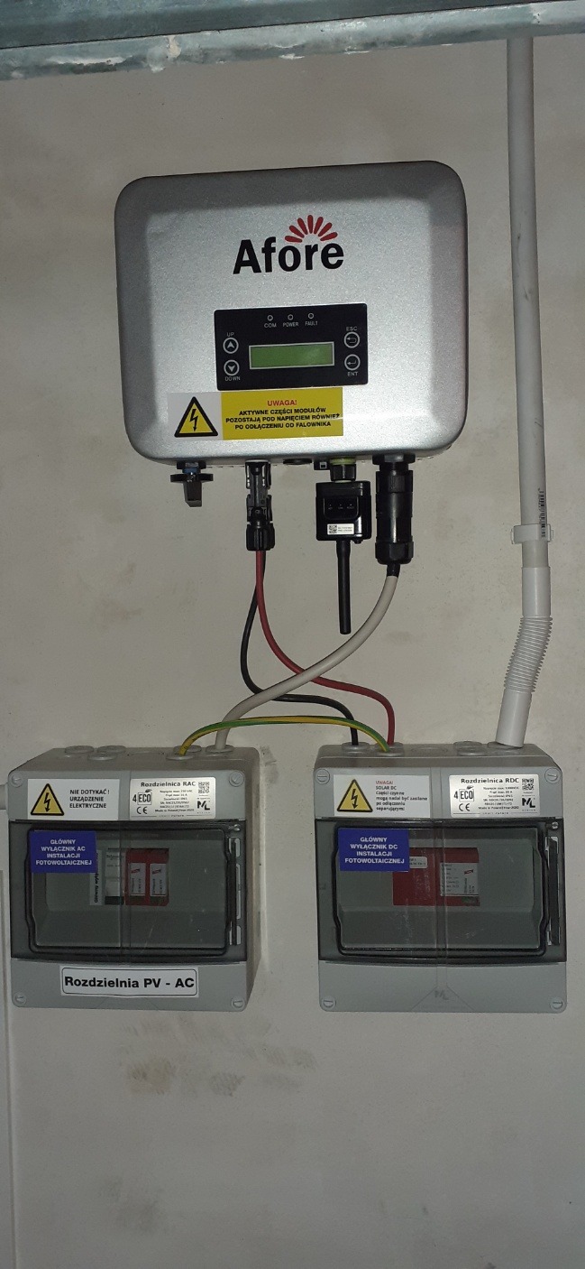 Falownik firmy Afore zamontowany na ścianie, poniżej dwie szare skrzynki rozdzielcze PV-AC i DC. Widoczne kable i oznaczenia ostrzegawcze o napięciu elektrycznym.