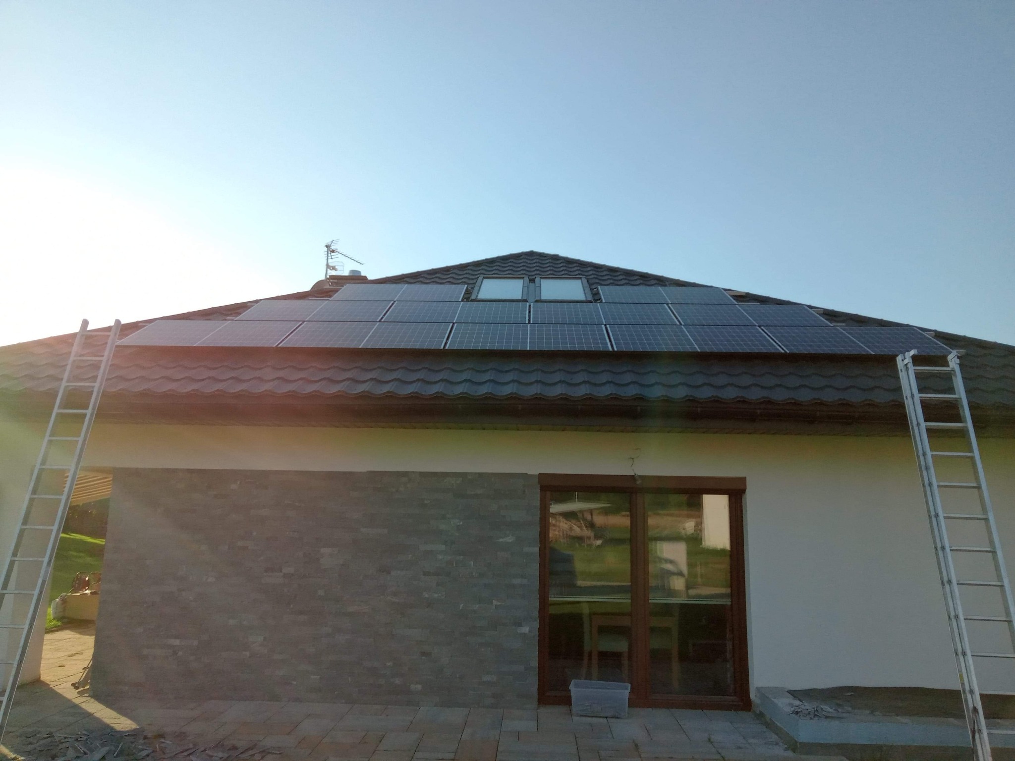 Panele słoneczne zainstalowane na dachu domu jednorodzinnego z dwoma oknami dachowymi, drabiny opierają się o ściany budynku, widoczne płytki chodnikowe i fragment elewacji z szarej cegły.