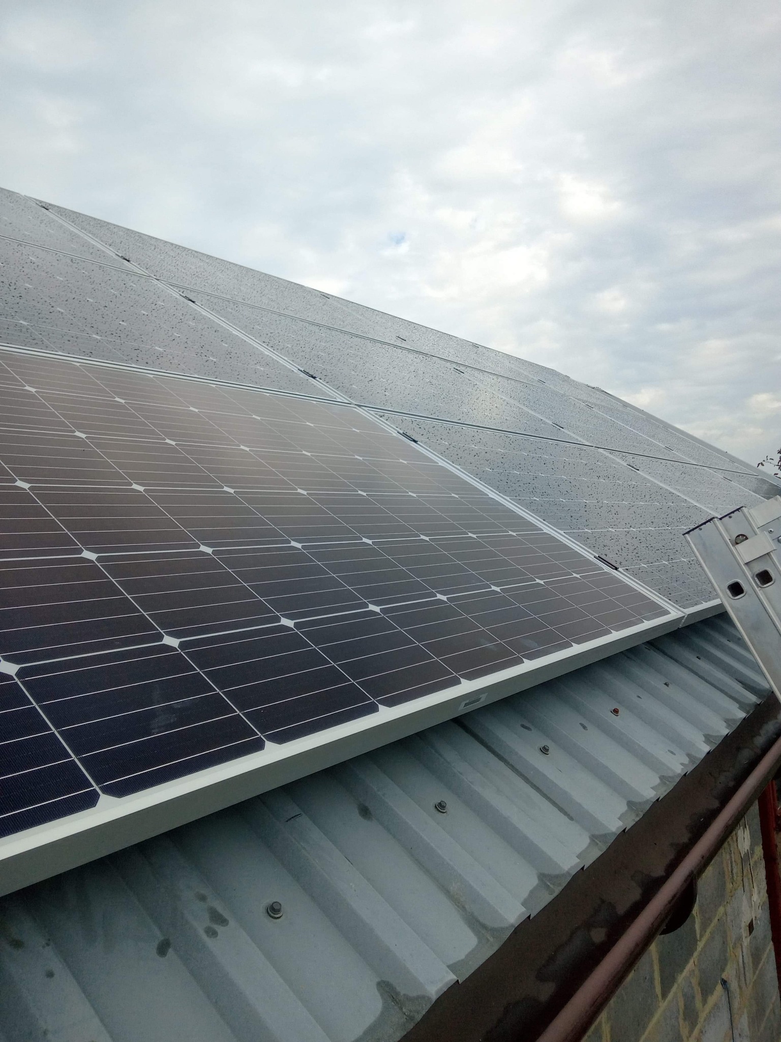 Panele słoneczne na metalowym dachu pokryte kroplami deszczu, drabina oparta o rynnę i ścianę budynku z cegły w tle pochmurnego nieba.