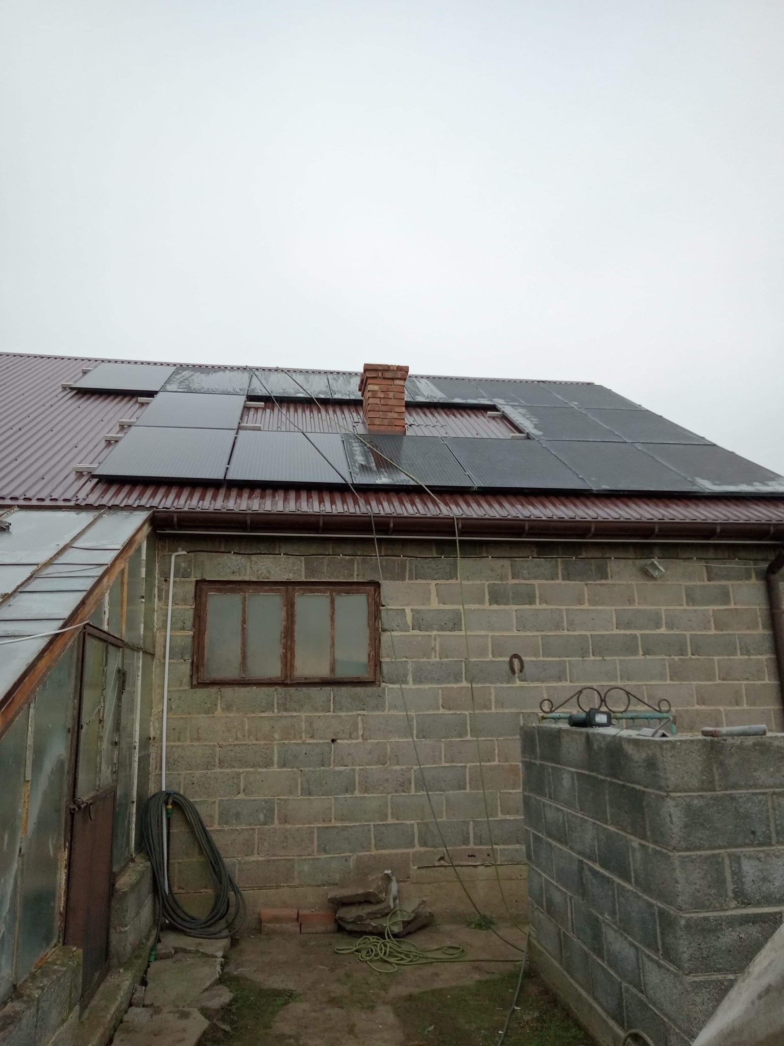 Instalacja paneli słonecznych na dachu budynku z cegły. Dach pokryty blachą trapezową w kolorze bordowym. Widoczny komin z cegły. Na ścianie budynku widoczne okno i zwinięty wąż ogrodowy.