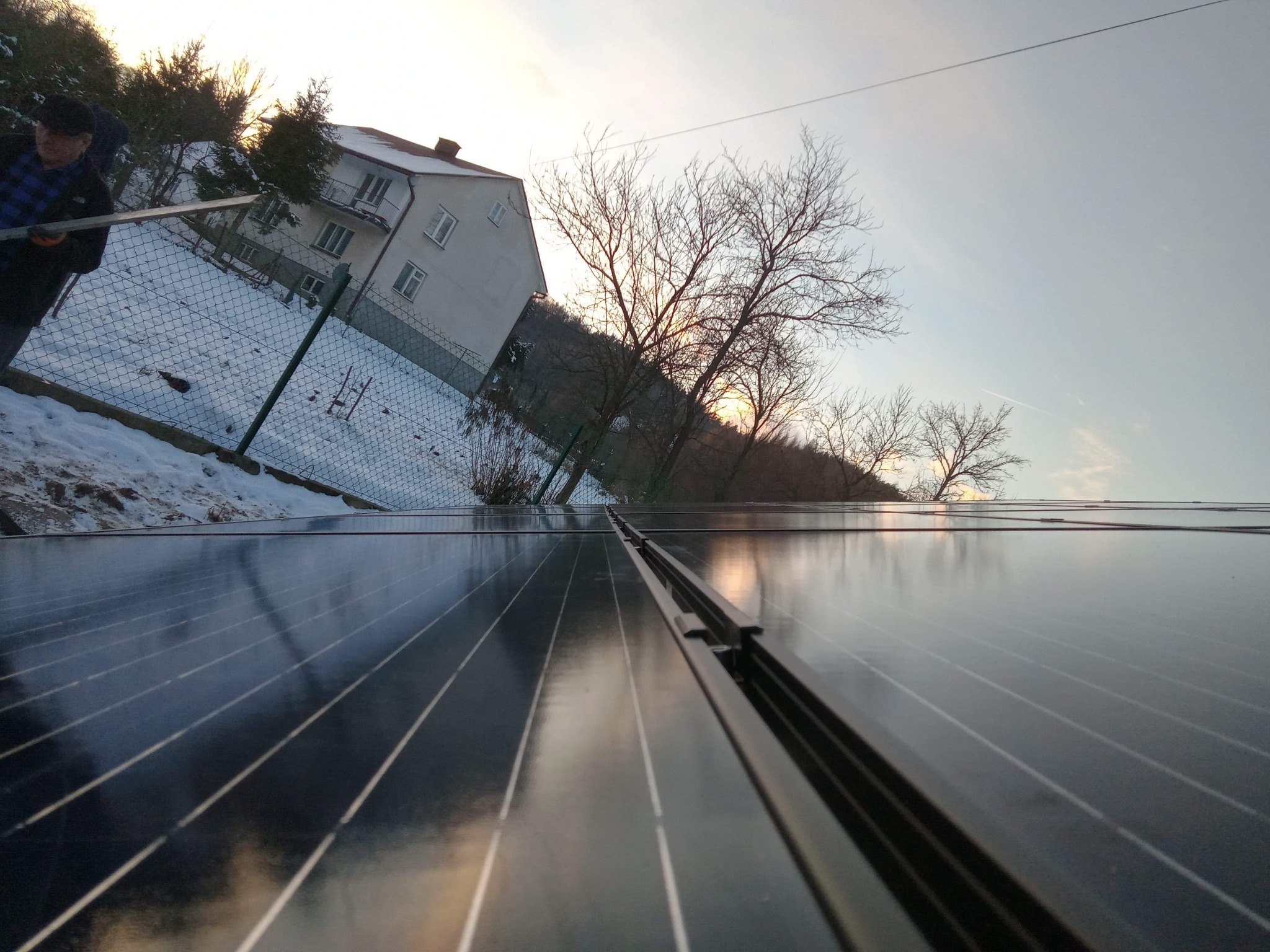 Montaż paneli fotowoltaicznych na dachu domu jednorodzinnego w zimowej scenerii, widoczny pracownik z narzędziem, odbicie nieba w panelach.