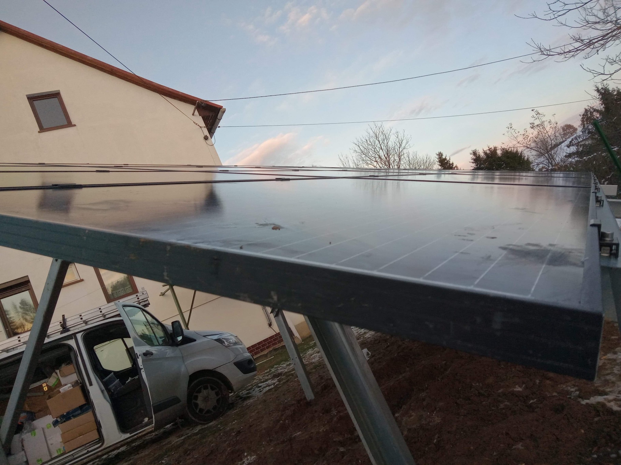 Instalacja paneli słonecznych w trakcie montażu, widoczny fragment dachu budynku, drabina oparta o ścianę i samochód dostawczy z otwartymi drzwiami wypełniony kartonami.