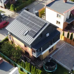 Widok z góry na dom jednorodzinny z panelami słonecznymi na metalowym dachu, widoczny fragment ulicy, ogrodu z trampoliną i zaparkowane samochody.