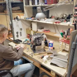 Serwisant w trakcie naprawy ekspresu do kawy na warsztacie, widoczne narzędzia i części urządzenia na blacie roboczym, podświetlone lampą warsztatową.