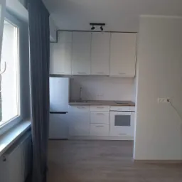 Jasna, nowocześnie urządzona kuchnia z białymi szafkami, sprzętami AGD i panelami podłogowymi, widok z perspektywy pokoju dziennego z zasłoniętym oknem po lewej stronie.