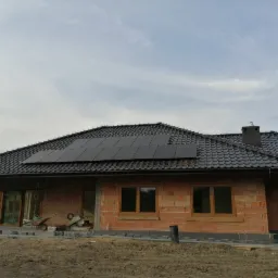 Częściowo wykończony dom jednorodzinny z zamontowanymi panelami słonecznymi na dachu z czarnej dachówki ceramicznej, widoczne okna z brązowymi ramami i fragment trawnika.