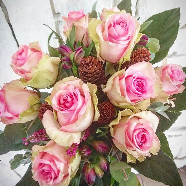 Delikatny bukiet z różowych róż o kremowych brzegach, uzupełniony drobnymi szyszkami i zielonymi liśćmi na tle białej, ceglanej ściany.