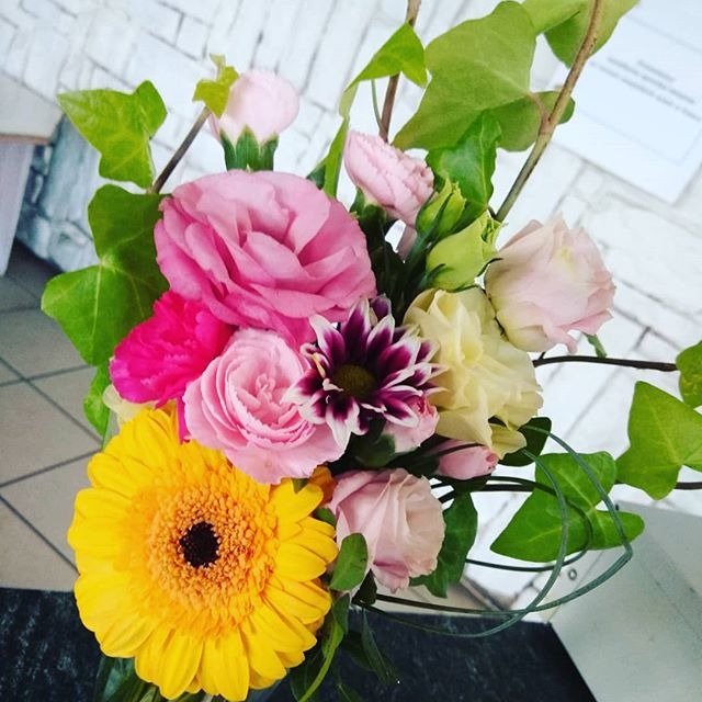 Kompozycja kwiatowa w odcieniach różu, żółci i fioletu z dodatkiem zielonych liści bluszczu, na tle białej ceglanej ściany.