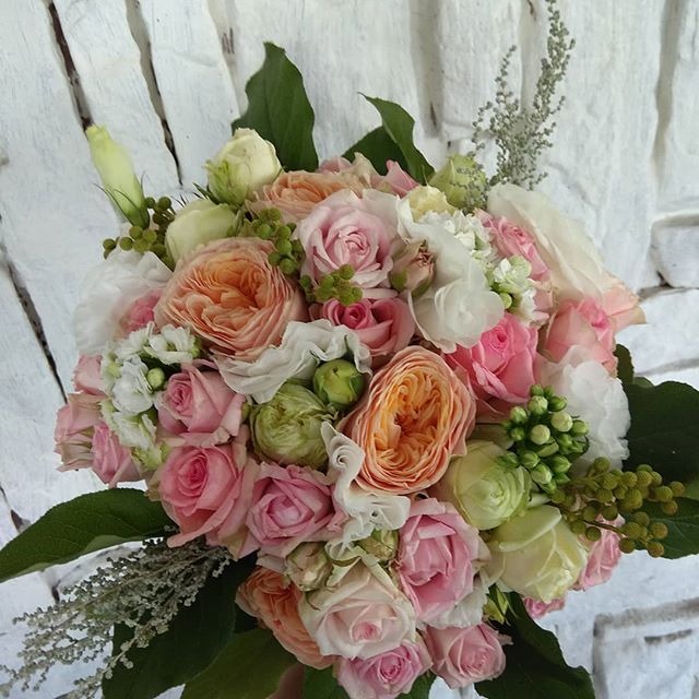 Delikatny bukiet ślubny z róż w odcieniach różu, kremu i brzoskwini, uzupełniony zielonymi dodatkami, na tle białej, ceglanej ściany.