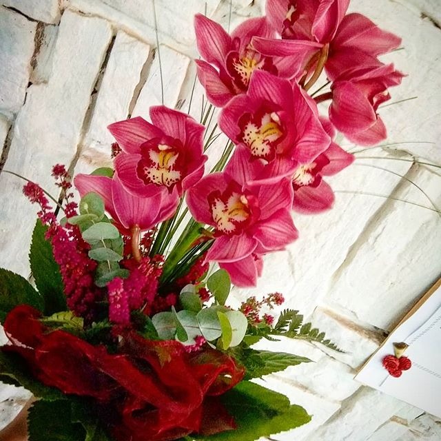 Kompozycja kwiatowa z różowych orchidei, drobnych czerwonych kwiatów i eukaliptusa, zawinięta w czerwoną siatkę, na tle białej, kamiennej ściany, z widocznym fragmentem białej kartki z małą...