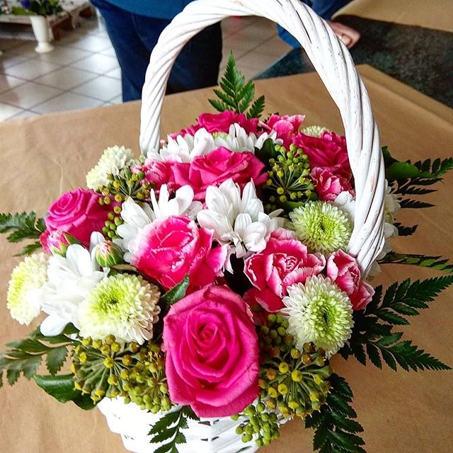 Kompozycja kwiatowa w białym, plecionym koszu z różowymi różami, białymi margaretkami i zielonymi chryzantemami na brązowym tle.