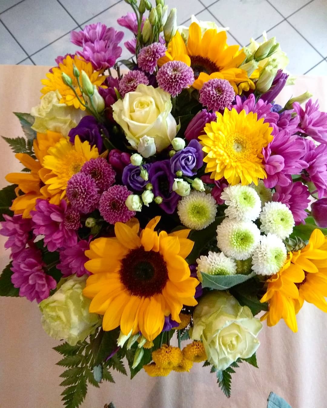Bujny bukiet z róż, słoneczników, gerber i chryzantem w odcieniach żółci, fioletu i bieli, ułożony na beżowym tle.