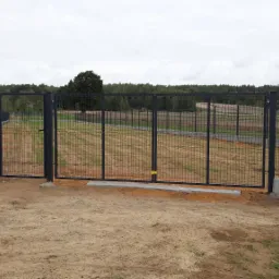 Nowoczesna, czarna brama wjazdowa z paneli ogrodzeniowych, osadzona na betonowej podmurówce, z widokiem na pole i las w tle.